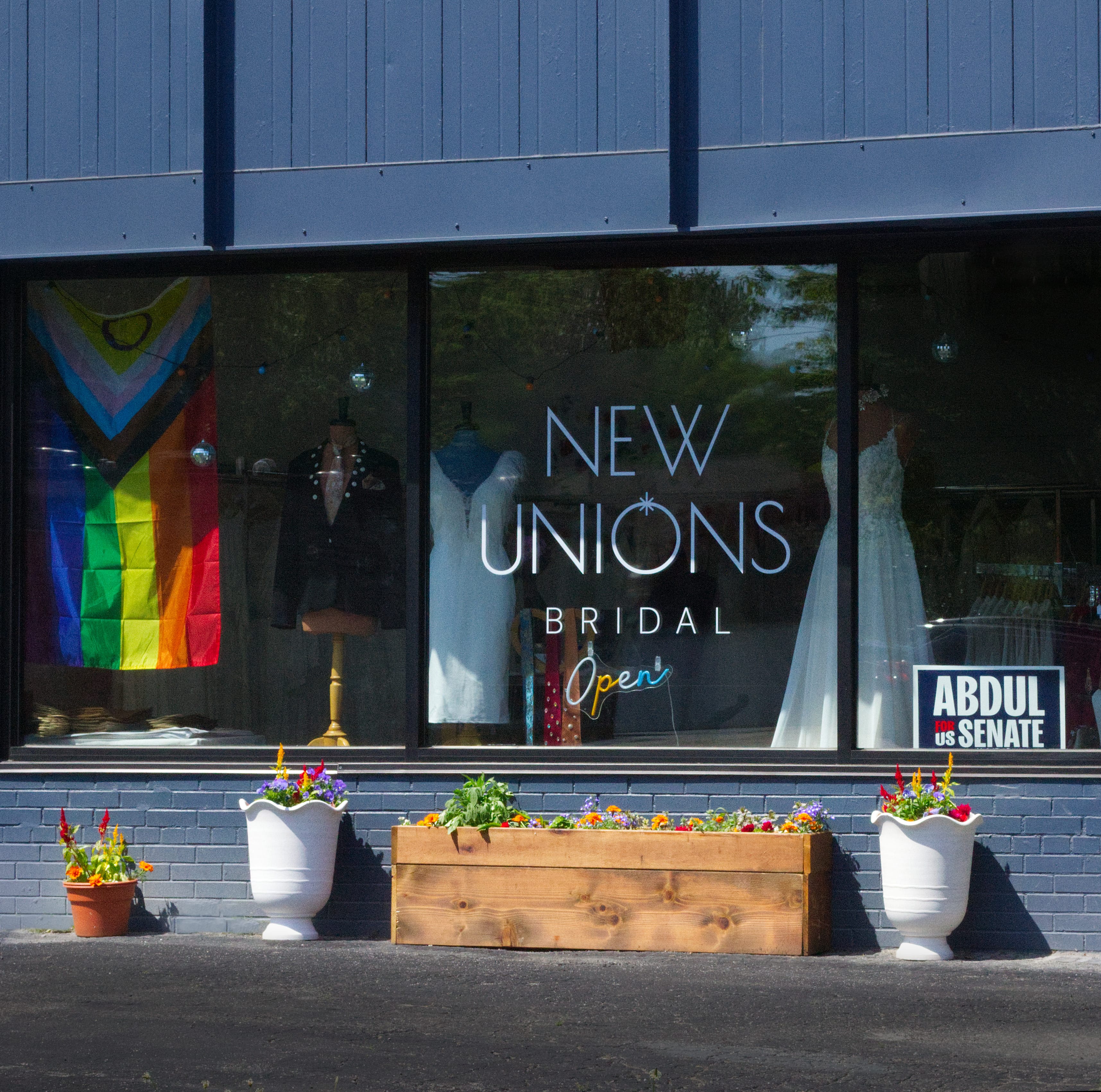 An exterior shot of the New Unions Bridal store from the street. The store has a pride flag and mannequins wearing wedding attire displayed in the windows, and a variety of potted plants outside.